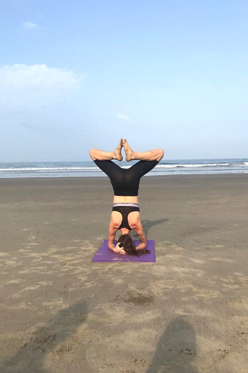 A yoga practitioner performing a headstand variation with controlled balance and core engagement. The pose showcases strength, focus, and alignment, with legs extended upward and hands providing support on the beach.